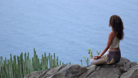 Amidst-island-mountains,-a-young-woman-practices-yoga-in-Lotus-position-on-a-rock-at-the-mountain's-summit,-overlooking-the-ocean